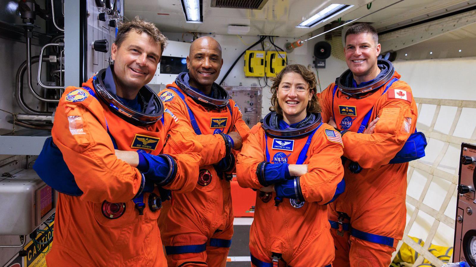 Three men and a woman in orange astronaut uniforms smile with their arms folded as they face the camera.