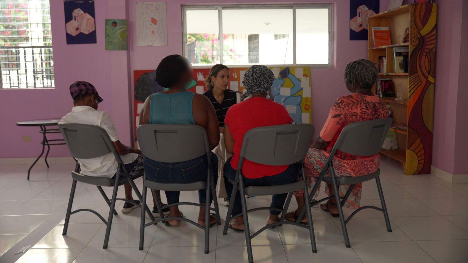Four women, sitting on grey plastic chairs, pictured from behind, speaking to BBC reporter Nawal Al-Maghafi. They are in a room with pink painted walls and a tiled floor.