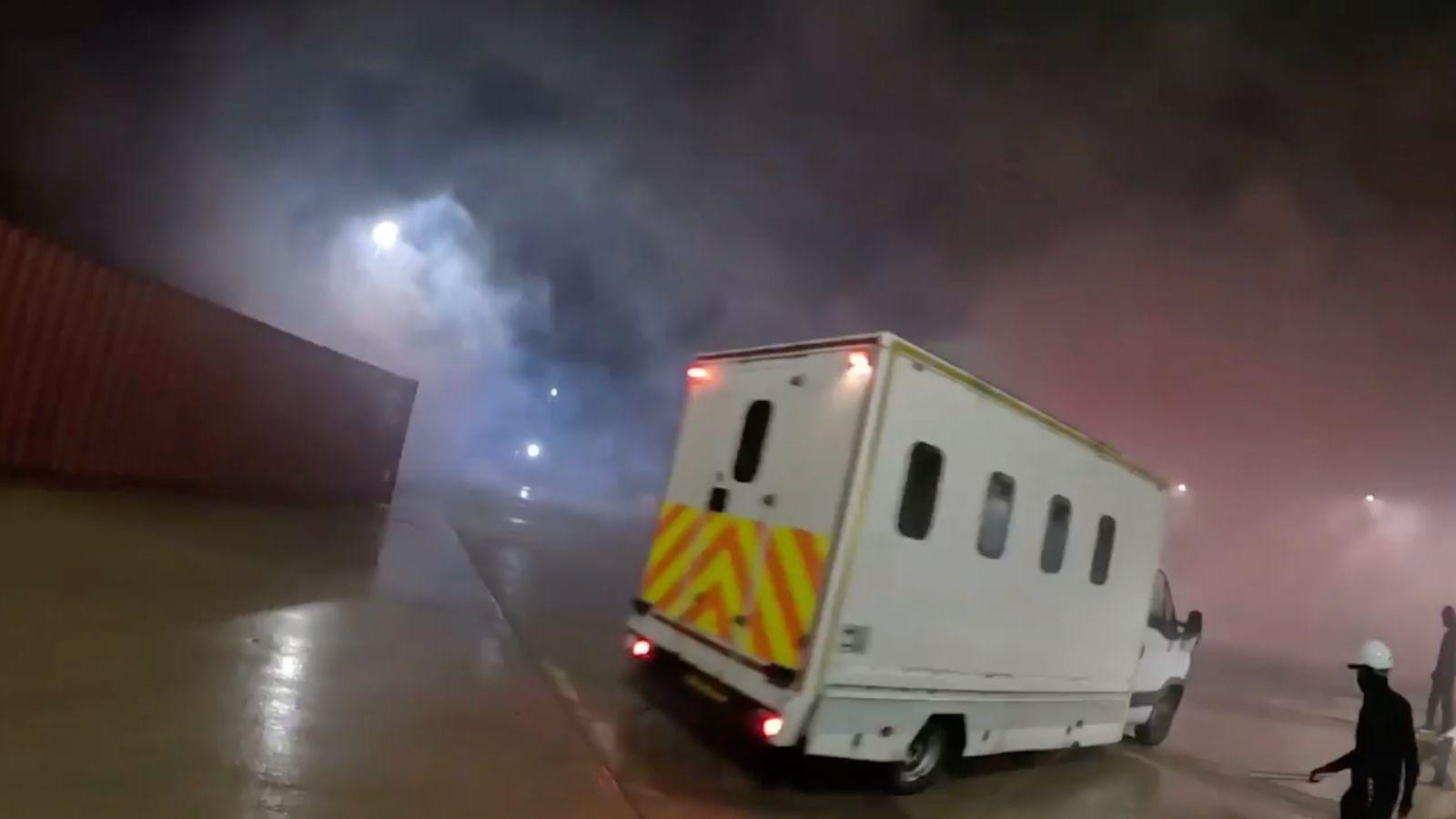 A white police van with yellow and orange hi-vis stickers on the back. It is being driven towards Elbit's warehouse building. There is lots of smoke in the air which is being illuminated by the red and blue floodlights on site. On the right there is a person wearing all black and a white hard hat. 