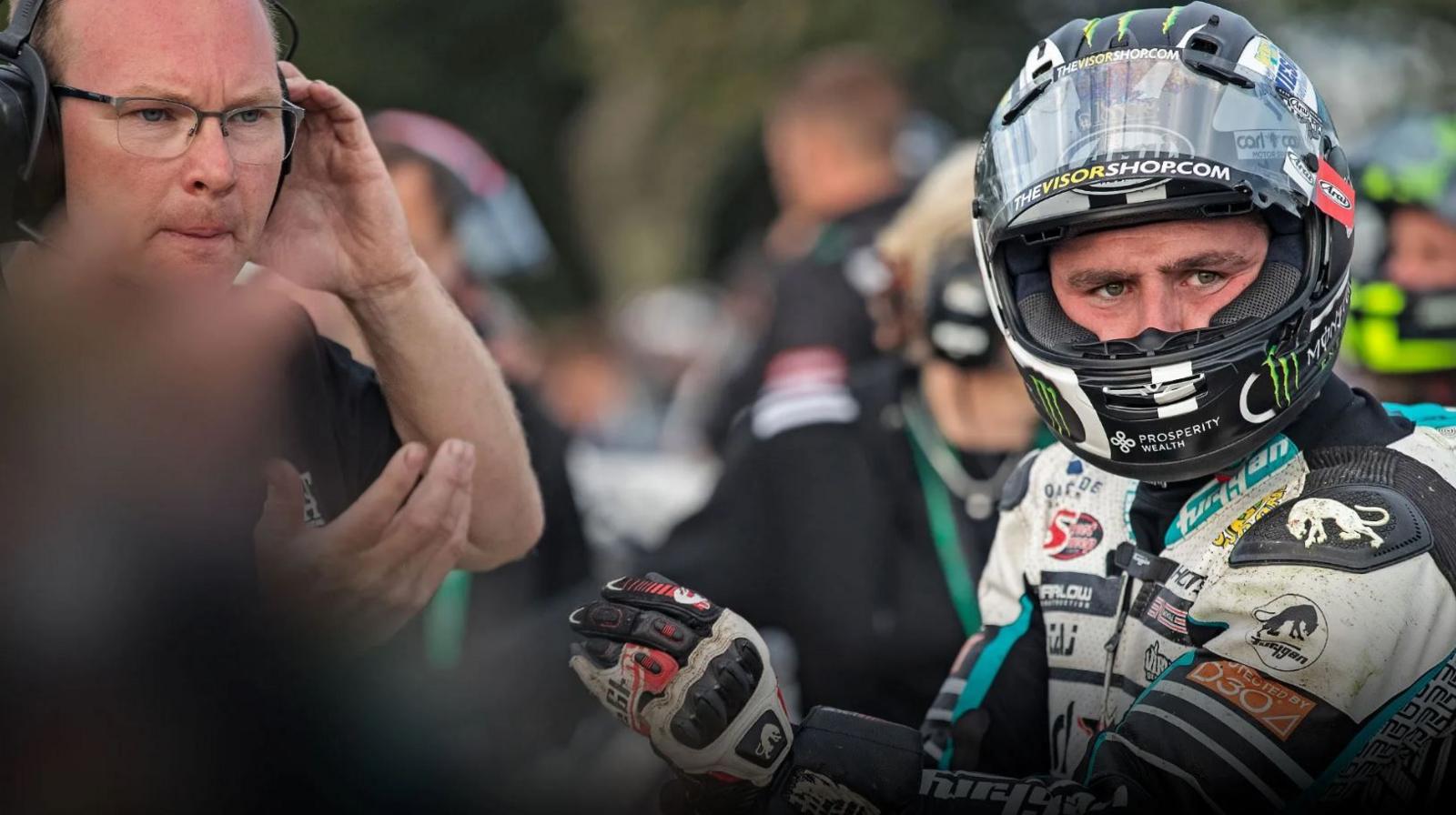 Michael Dunlop, who is in his leathers and has his helmet on. The visor of the helmet is up. Richard 'Milky' Quayle is standing to his left beckoning someone else over.