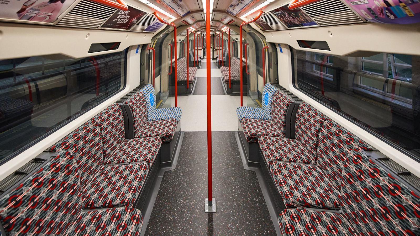 The inside of a Central Line tube carriage. Patterned red and blue seating runs along either side of the carriage, with windows above showing darkness outside. Red poles run from floor to ceiling in the middle of the carriage seating area, and in the standing areas by the doors.