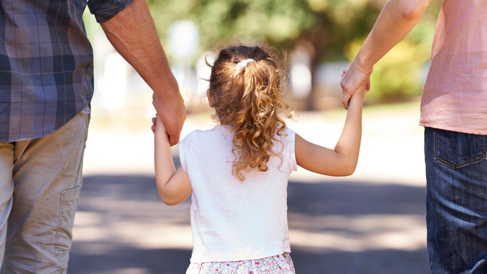 Young girl standing in the middle of her mother and father, holding both of their hands. They're all facing away from the camera.