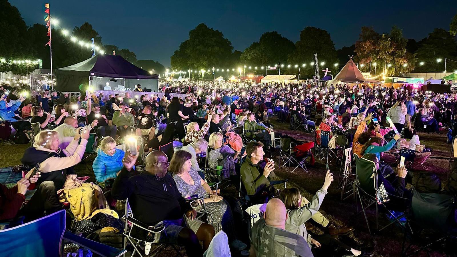 A crowd of people, sat on camping chairs, at Darley Park concert at night with a few tents and lighting in the background 