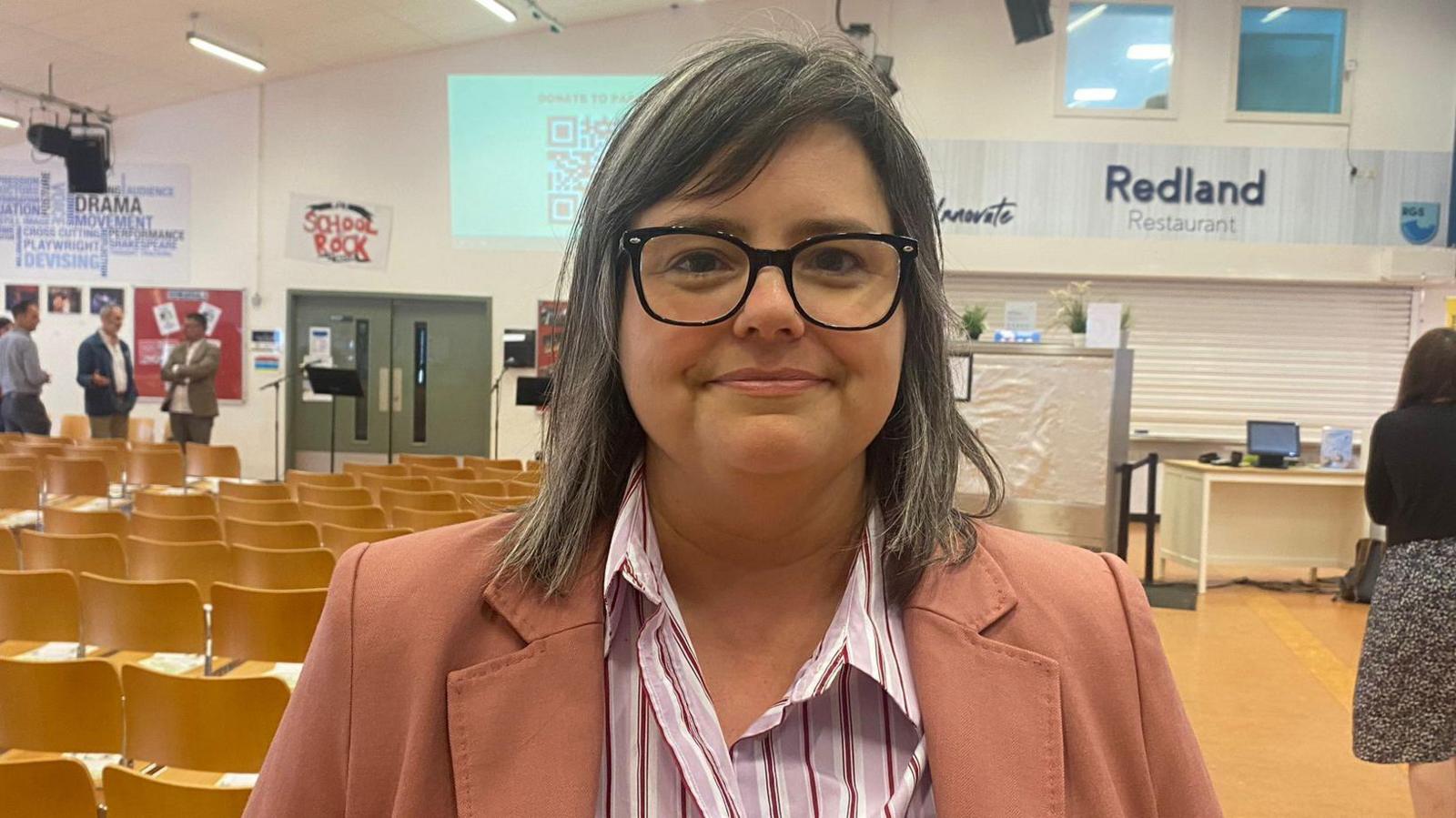 Melanie Sweet standing in a school hall. She has medium length straight, grey hair and is wearing thick-rimmed black glasses, a pink and white striped shirt and a salmon pink blazer.