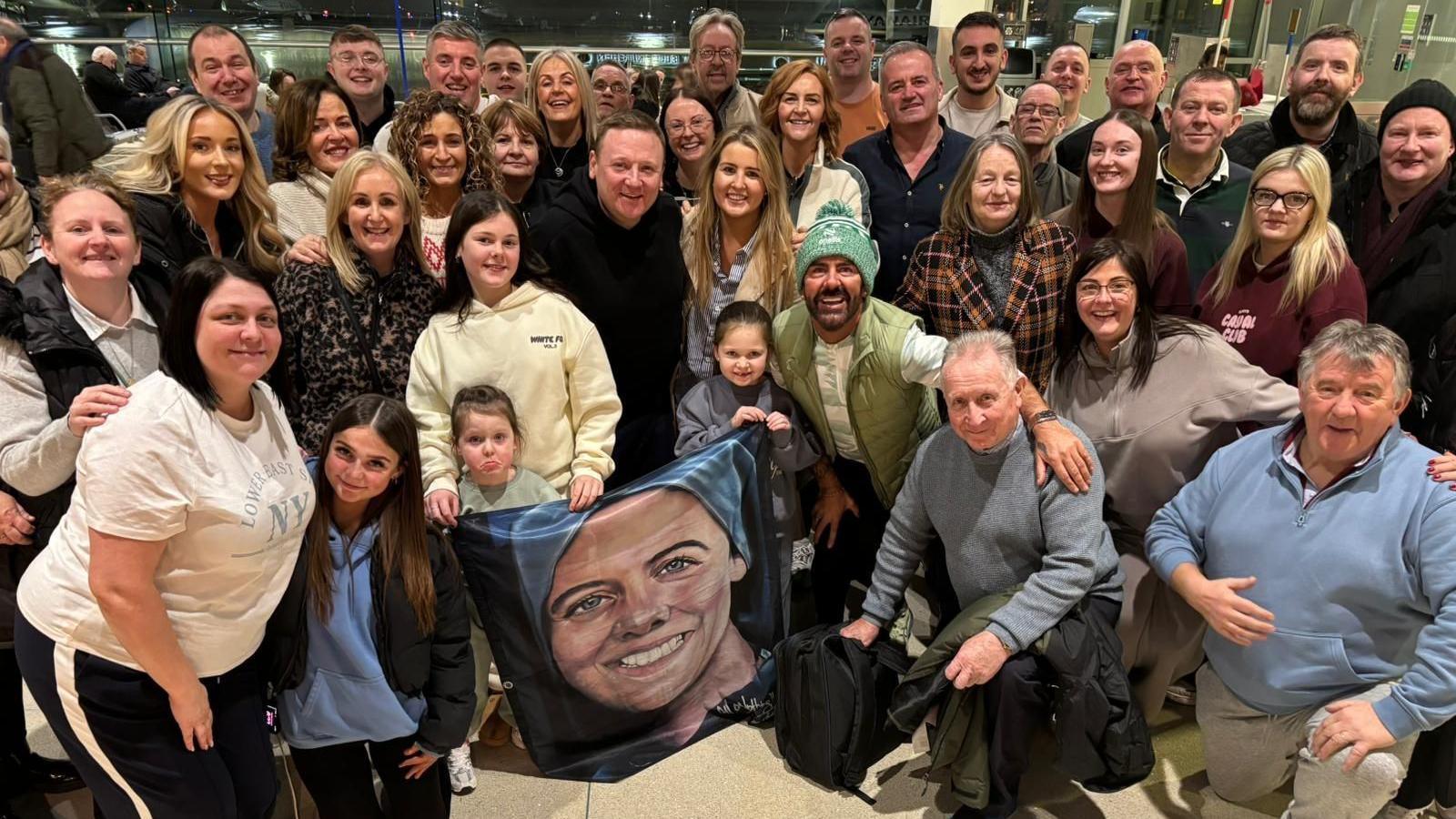 A large group of people all gather around while smiling at the camera. A few people in the front row are holding up a flag with a portrait of Sr Clare on it.