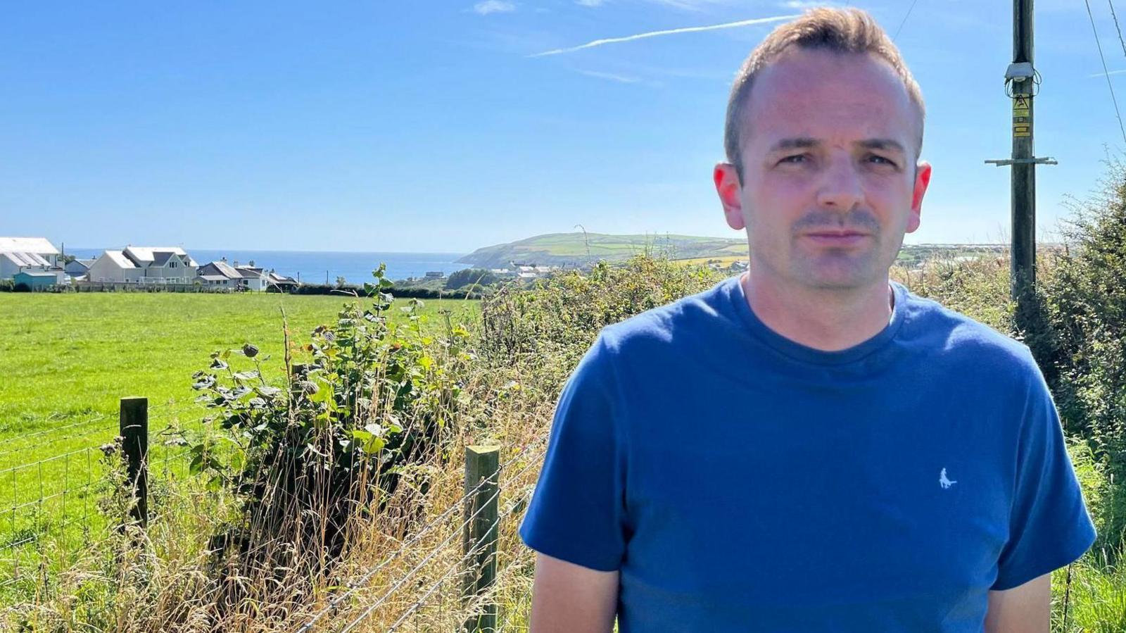 Gareth Williams, wearing a blue T-shirt, standing near a field with homes and the sea in the background