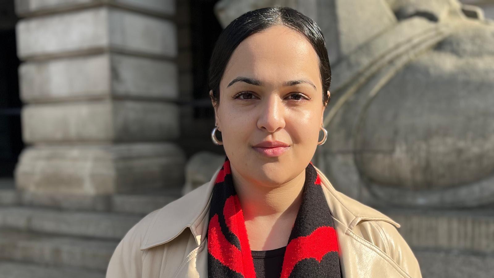 Nadia Whittome, MP for Nottingham East,  stood outside, with Nottingham council house behind her. She has dark hair which is scrapped back and is wearing gold earrings, as well as a red and black scarf. 