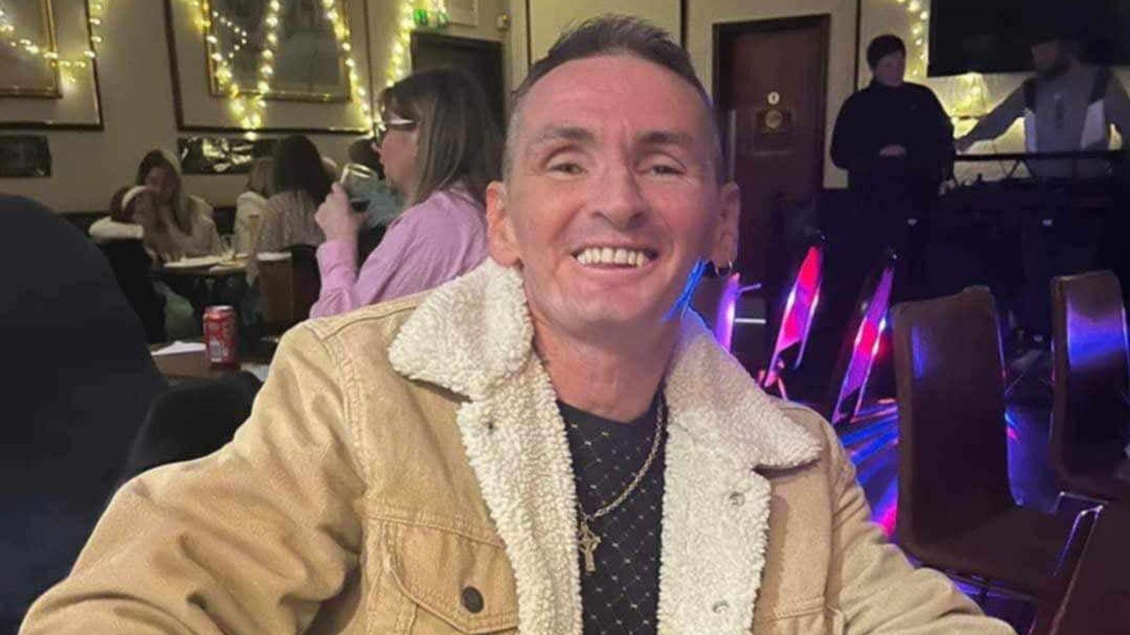 A man with a tan jacket, black shirt and gold chain around his neck smiles for the camera. He has short cropped hair and is sitting down, in what looks like a bar or community event, with other people sitting at tables behind him. 