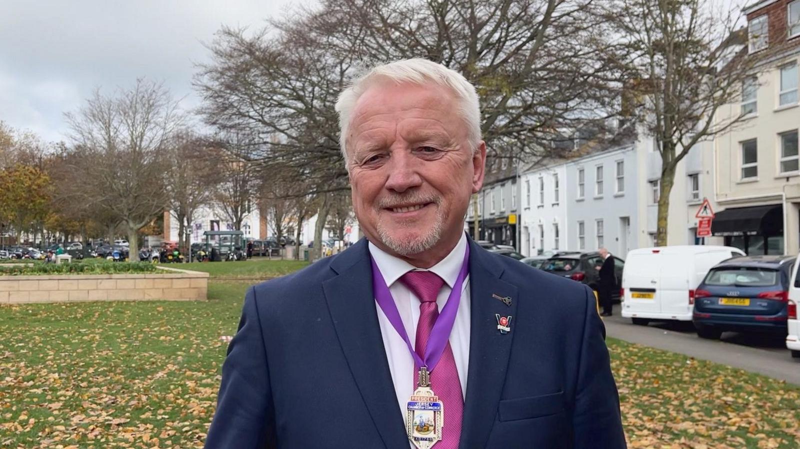 Lee Madden, a man with short white hair and a goatee-style beard. He is wearing a white shirt, dark blue suit and a pink tie. He has a poppy appeal badge on his lapel, and a medal with a purple ribbon around his neck. Behind him, cars and buildings are visible on the right of the image, while fallen leaves lie on the grass to the left. There are a number of trees behind his head.