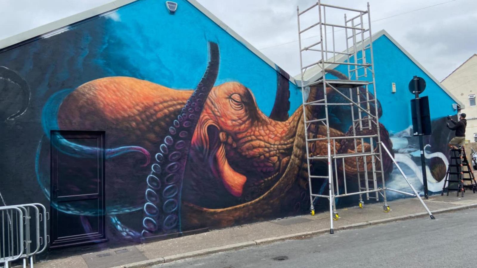 A mural of an octopus in the sea drawn on the side of a building, with the artist painting from a stepladder.