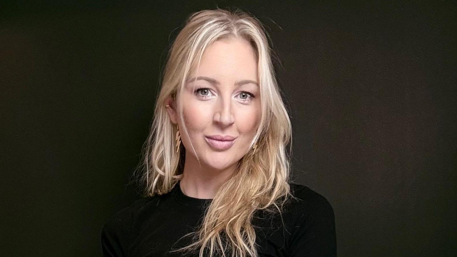 Liz is wearing a black top and is stood against a black background. She has long blonde hair and is wearing gold-coloured earings.