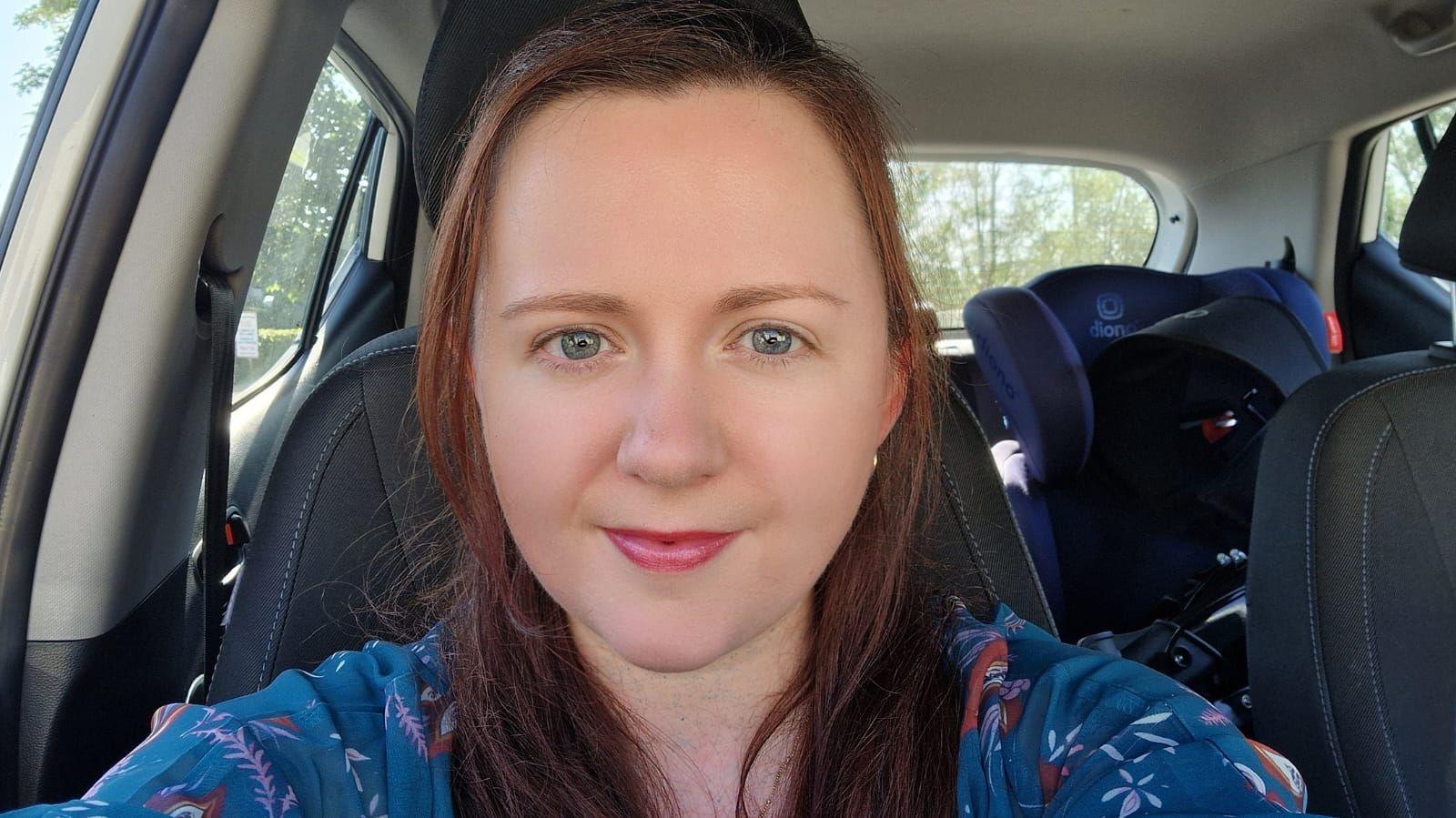 Rachel has taken a selfie photo in her car. She has shoulder length brunette hair and is wearing a blue blouse top.