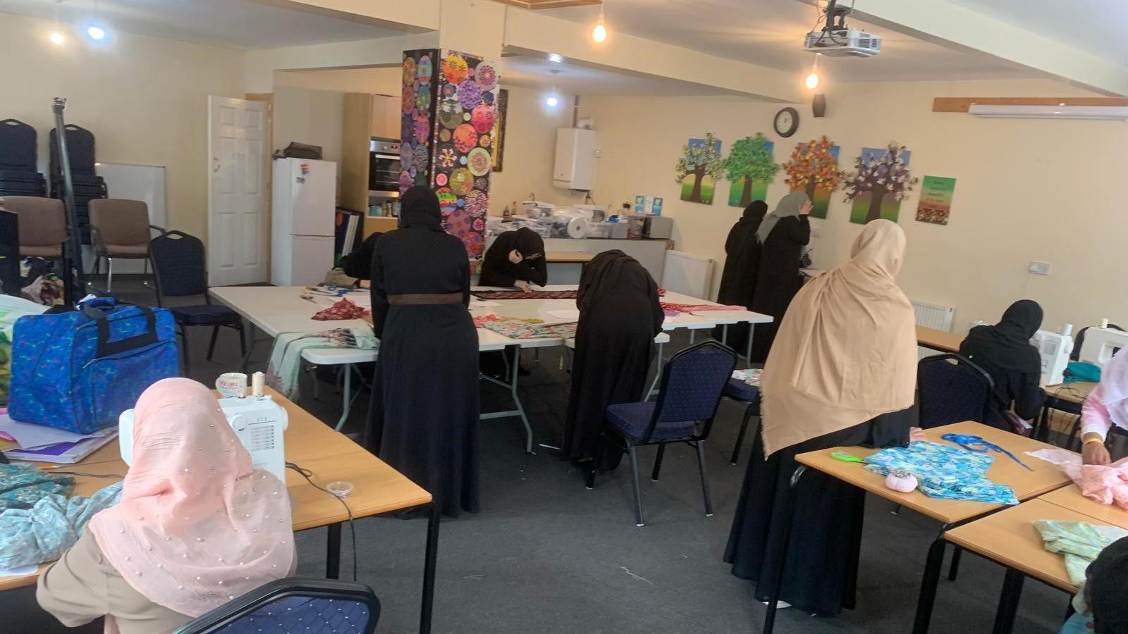 Nine women in headscarves sit at tables sewing or cutting out material