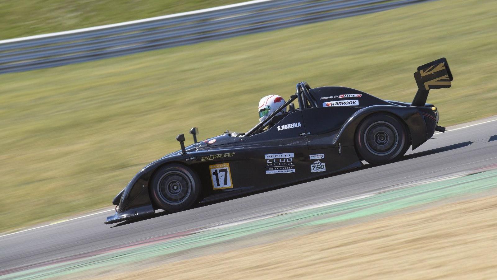Stephanie Hobeika wearing a helmet in a car, racing on a track at high speed. The car is branded and is a dark colour