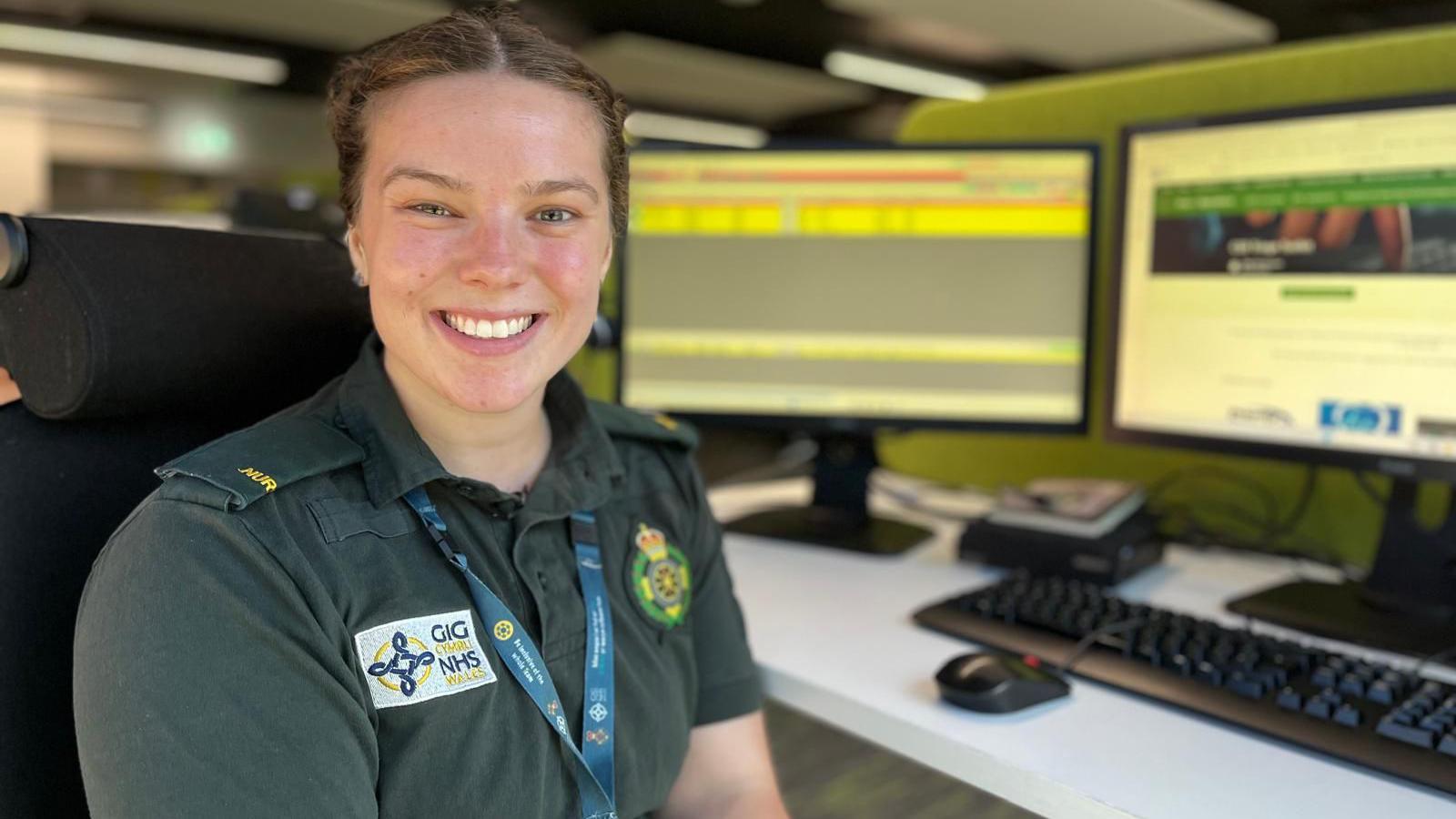 Ruth is wearing the green uniform of the ambulance service, with embroidered logos on it and a staff lanyard. She is sat at her desk with screens behind her. She is smiling at the camera.