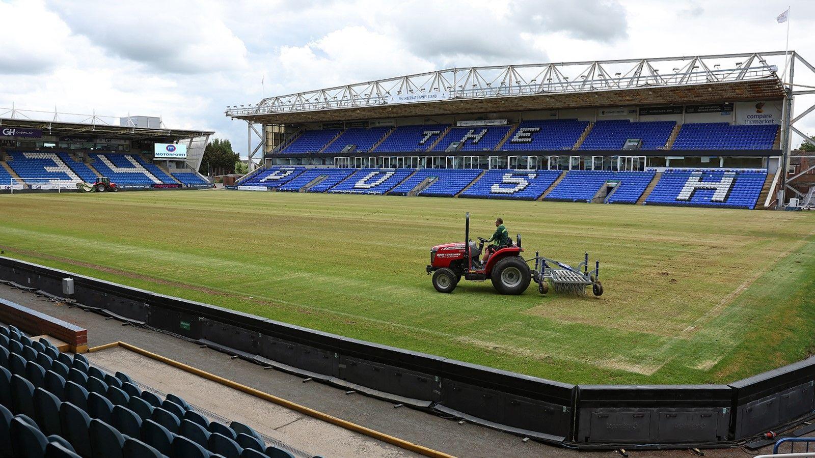 A view of Peterborough United's stadium, with a man riding a small red tractor across the pitch. The tractor has a rake attachment at the back. The stand opposite has blue seating, with The Posh spelt out in white seating.