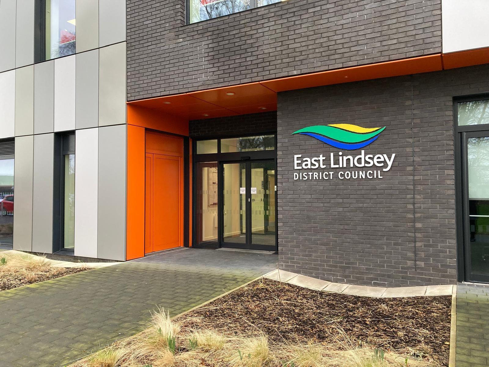 A photograph taken outside the main doors of East Lindsey District Council showing the council's logo. The building is grey, with a bright orange panel to the left of the main entrance.