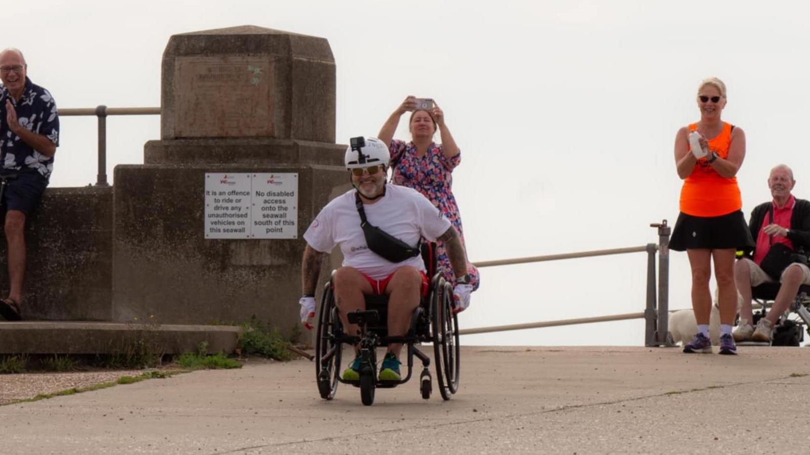 Ben Parker wearing a white t-shirt and red shorts travelling in his wheelchair, he is smiling. People are standing behind clapping him and taking photos.