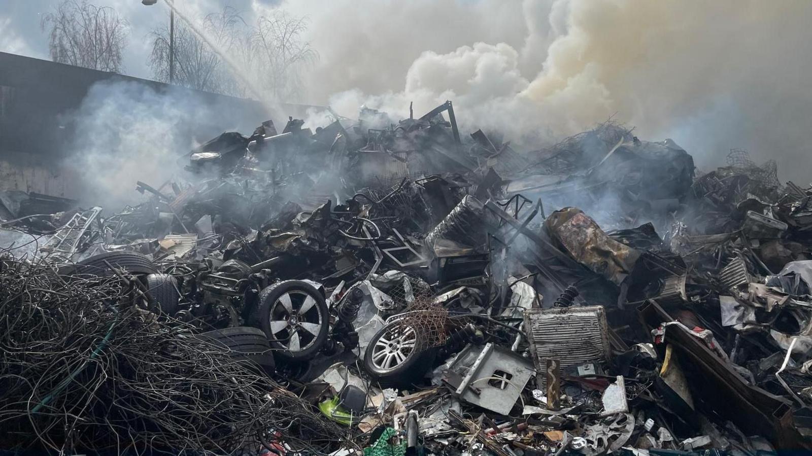 An aerial hose targets water down on a mound of tyres and other burning debris with smoke rising from it