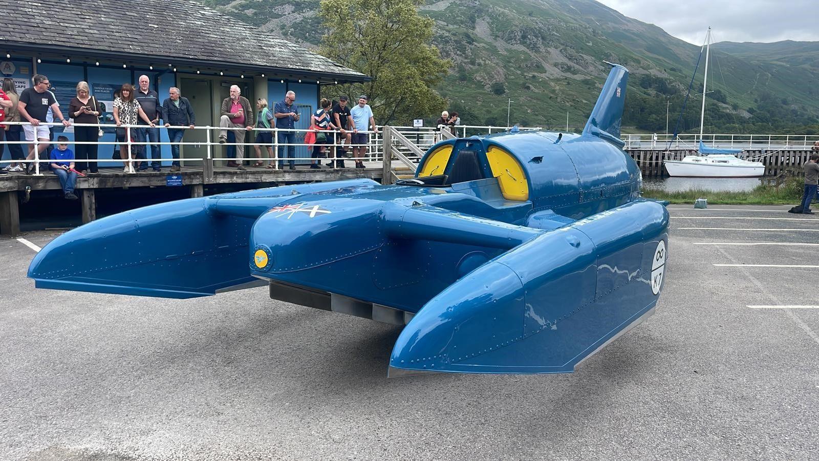 Bluebird - a jet-powered hydroplane - on display at Glenridding Pier in a car park. A number of people are looking on.