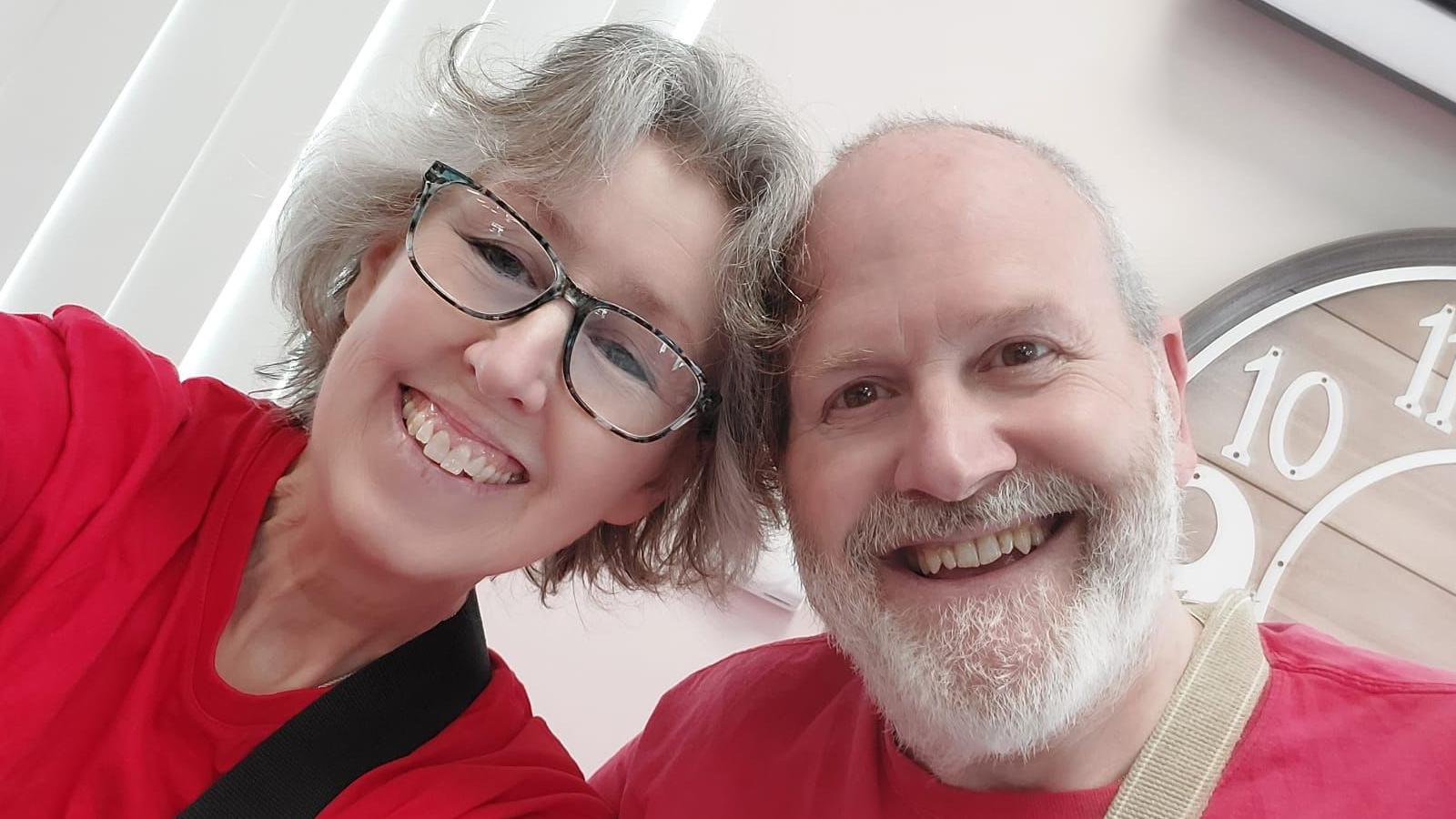 Roz Hall in a red t-shirt and wearing glasses next to Graham Hall. They are both smiling and leaning into each other.