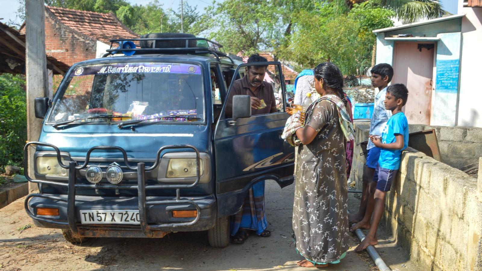 A man wearing a brown shirt and blue cloth tied around his waist hands cash to a female customer who is wearing a grey night dress. The man stands alongside his blue van. 