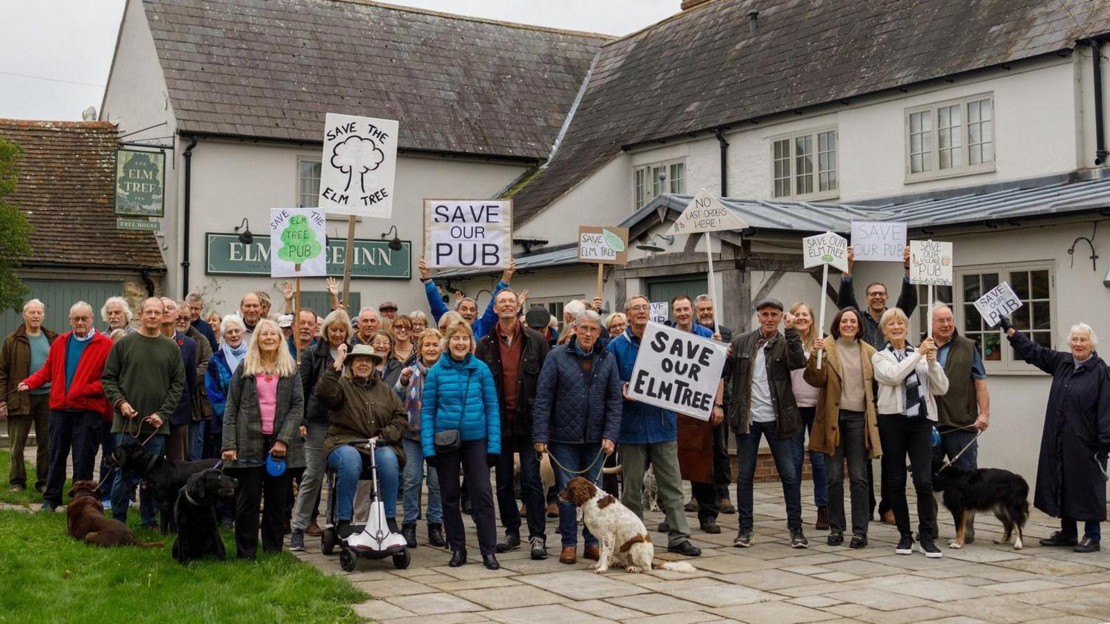 About 50 people and some dogs standing in front of the Elm Tree pub. Many are holding up signs saying 'save our pub' and 'save the Elm Tree'.