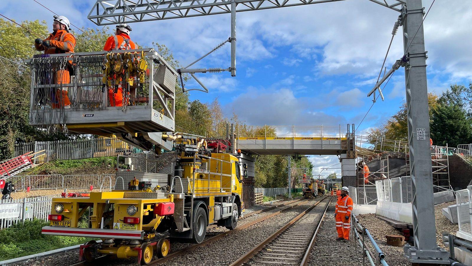 Engineers are standing by a railway track dressed in orange outfits and hard hats. A further two engineers are using a platform to reach overhead power lines.