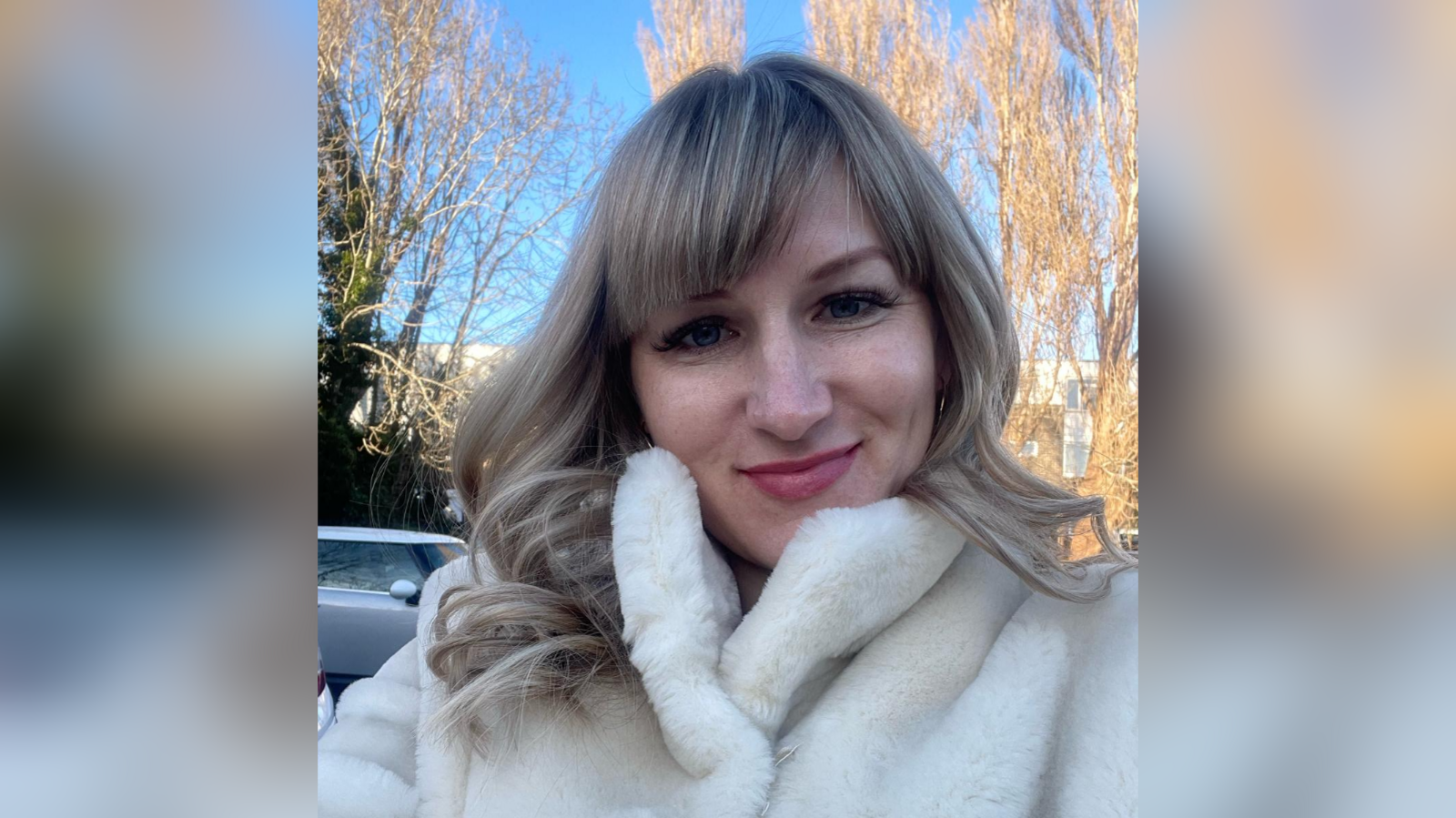 A woman smiles in a white fluffy coat. She has blonde hair and a fringe. In the background there are trees and a car. 