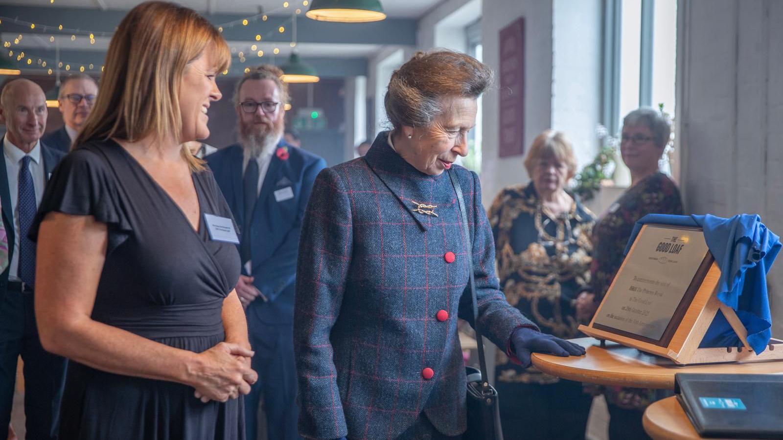 Princess Anne wearing a navy jacket with pink buttons looking at a wooden plaque sitting on a table surrounded by bakery staff. 