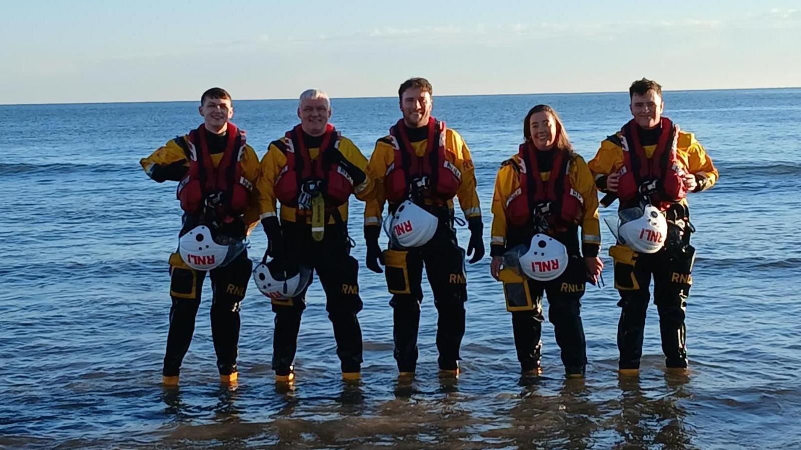 Five Sunderland RNLI crew smiling as the stand in the sea. Their clothing is black and yellow, Their white helmets are attached to their belts. 