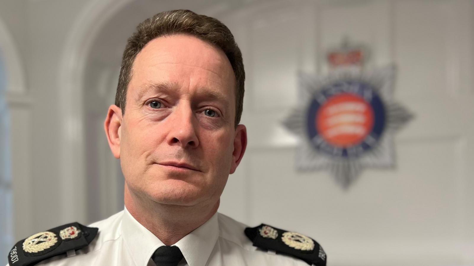 Essex Police chief constable BJ Harrington standing in front of the Essex Police badge. He is dressed in uniform and has a pained expression of his face.