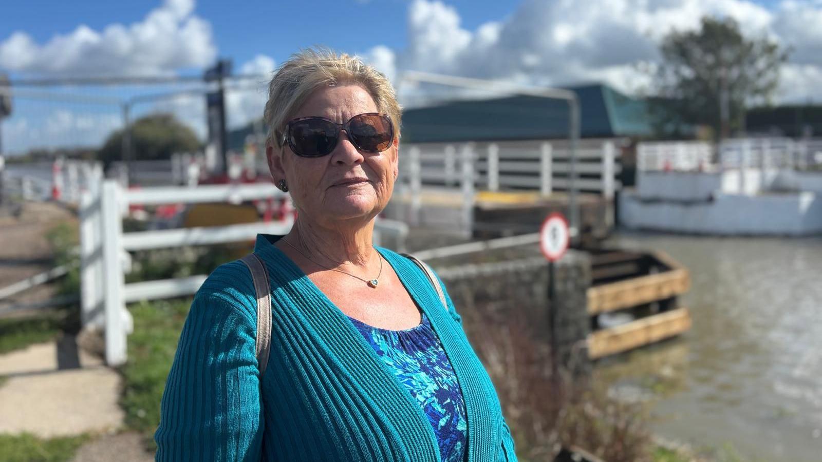 A woman with short blonde hair wearing sunglasses, a turquoise cardigan and blue top stands in front of the canal and bridge, looking at the camera