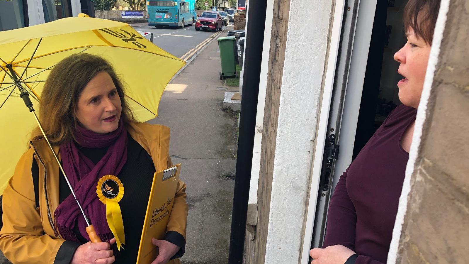 A picture of Lara Pringle canvassing. She is talking, on a doorstep, with a woman who has dark hair and a brown top. Ms Pringle has a yellow umbrella and is wearing a yellow coat and a purple scarf with a Lib Dem rosette.