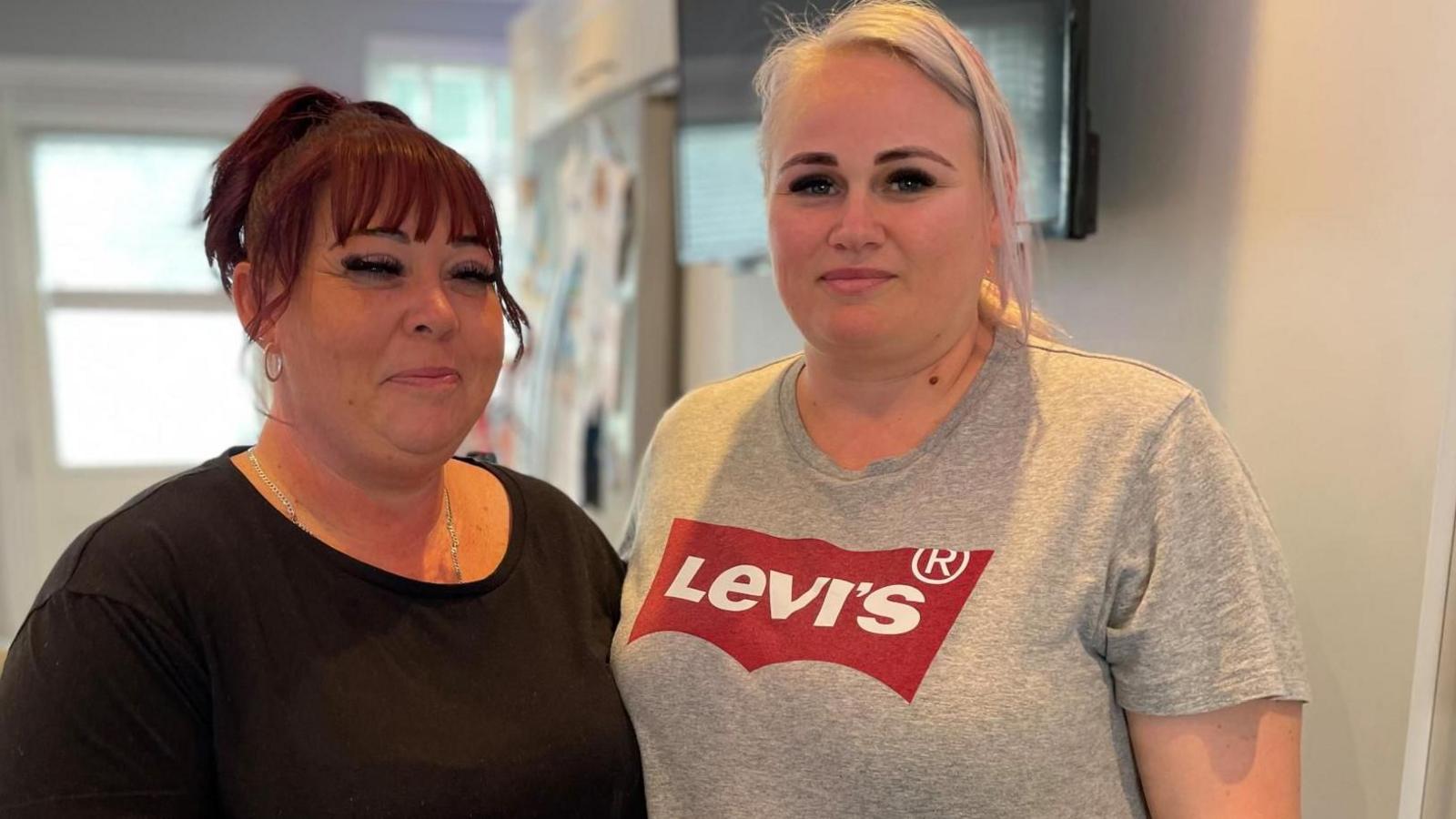 Two women stood together pose for the camera. On the right, a blonde woman with a grey tshirt. On the left a woman with dark red hair and black top.