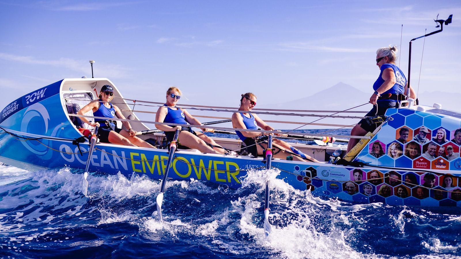 A rowing boat with three women using the oars and another woman at the back of the boat who is the cox. The waves are crashing against the side of the boat and the sky is a pale blue in the background. 