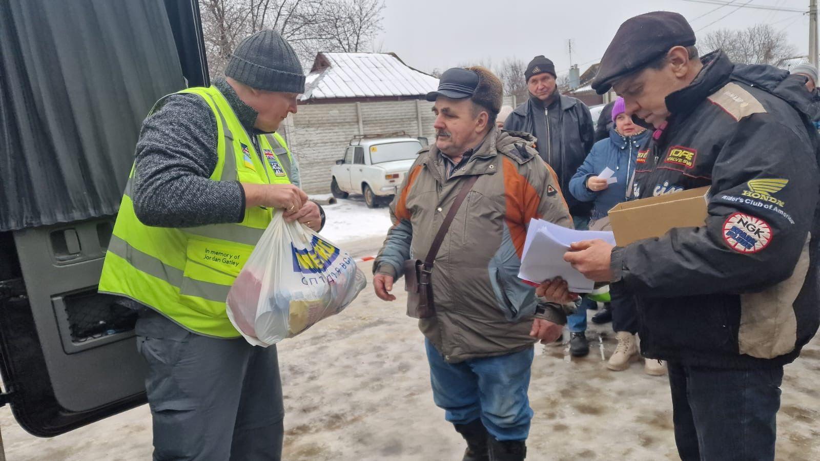 A man wearing a woolly hat, a long-sleeved top, grey trousers and a hi-vis vest hands out a bag of supplies to a waiting man.