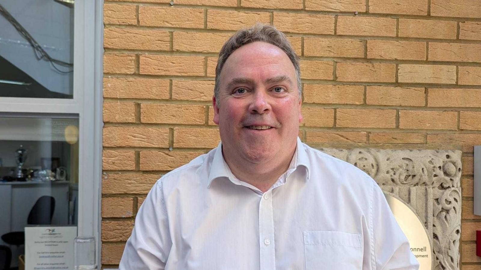 Mayor Jason Perry smiles to camera wearing a white shirt. He is standing outside beside a brick wall