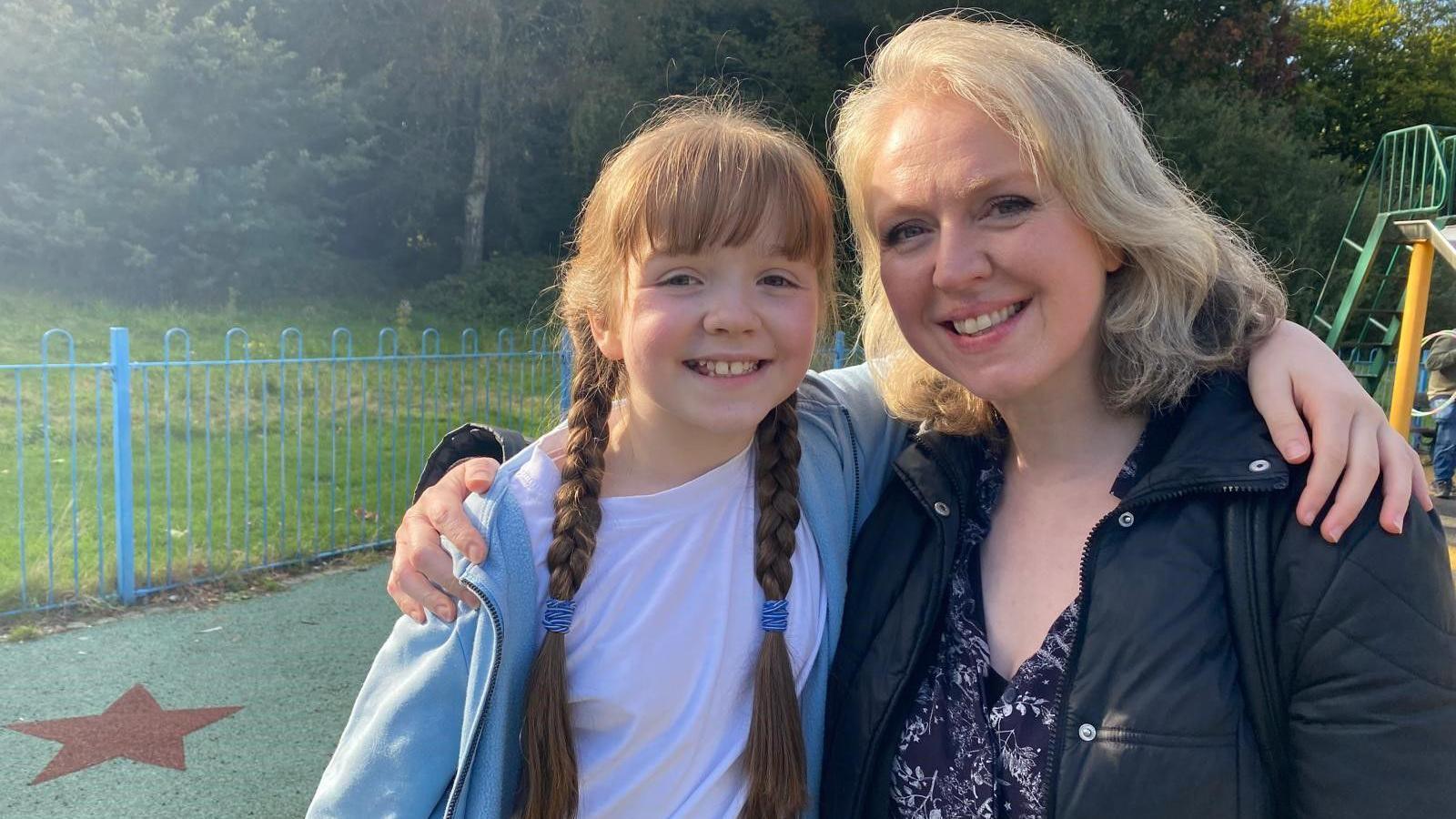 Phoebe, 10, and her mother Jemma sit on play equipment in the park. They are smiling and have their arms around each other. 