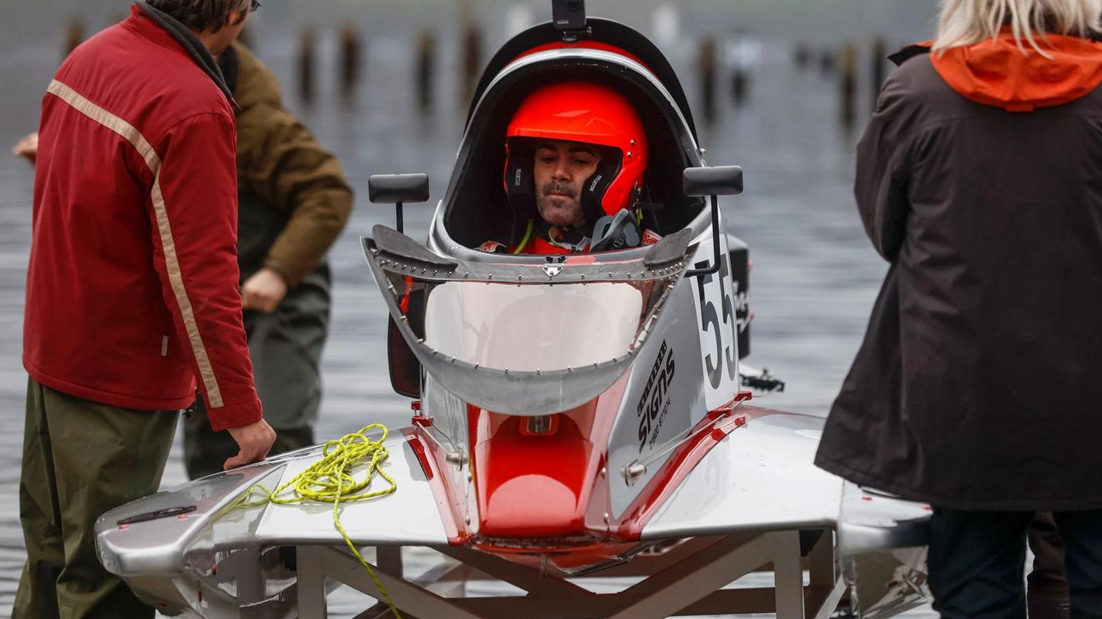 A man in a red helmet is pictured sitting inside a white and red powerboat near the water's edge. The boat has the number 55 on it. There is a blonde-haired woman to the right of the boat and two men to the left. The man in the boat is looking nervous. The boat is curved and has wings. 