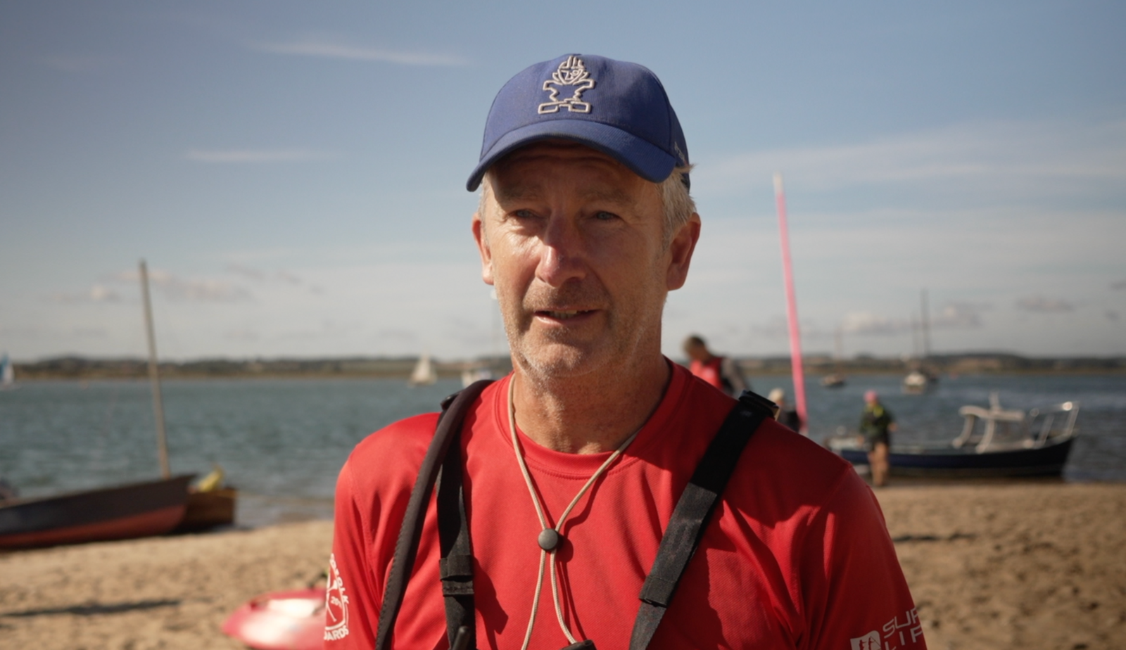 Andy Holland, one of Charlie's sponsors, talking to Look East on the beach, wearing a red t-shirt and blue cap with multiple boats on the water behind.