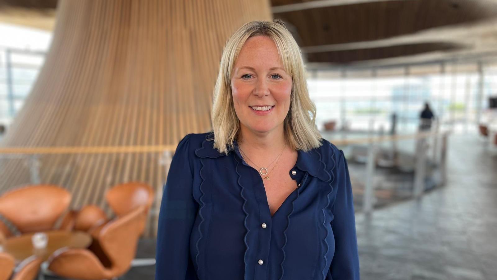 The image shows Nerys Evans standing in the Senedd in front of the funnel. She is looking at the camera and smiling. She is wearing a navy top.