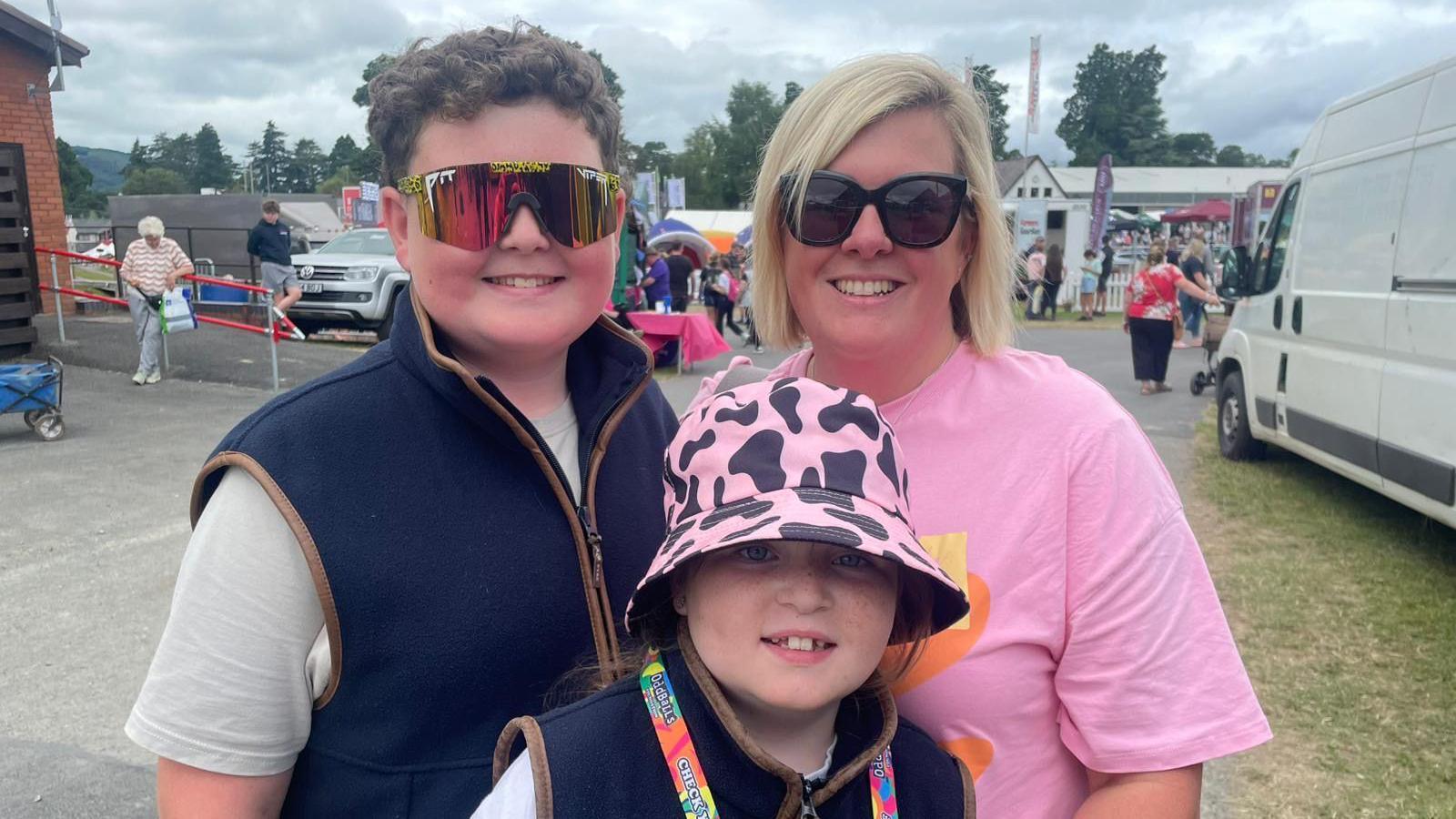 Ella smiling with her two children at the show with stalls behind her. Her son is standing to the left of her and is wearing large colourful sunglasses, a blue gilet and a grey t-shirt. He has short curly brown hair. Ella's daughter is standing in front of her. She is wearing a pink and black bucket hat with a blue gilet. Ella is wearing a pink t-shirt and black sunglasses. She has short blonde hair.