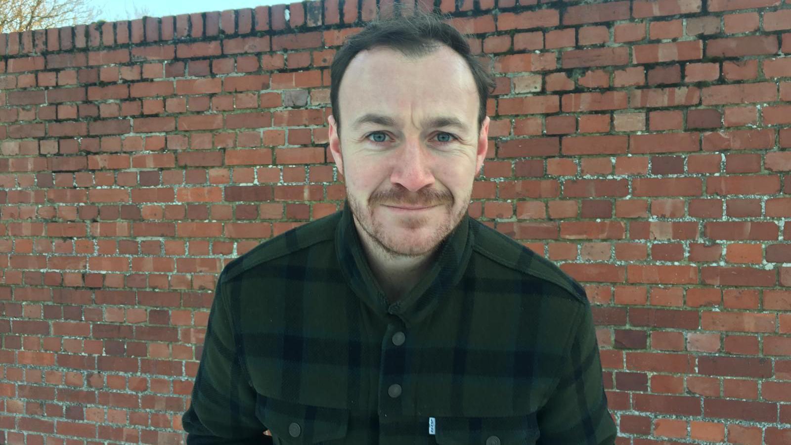 Matt Aitken is standing in front of a red brick wall looking at the camera. He is wearing a checked shirt.