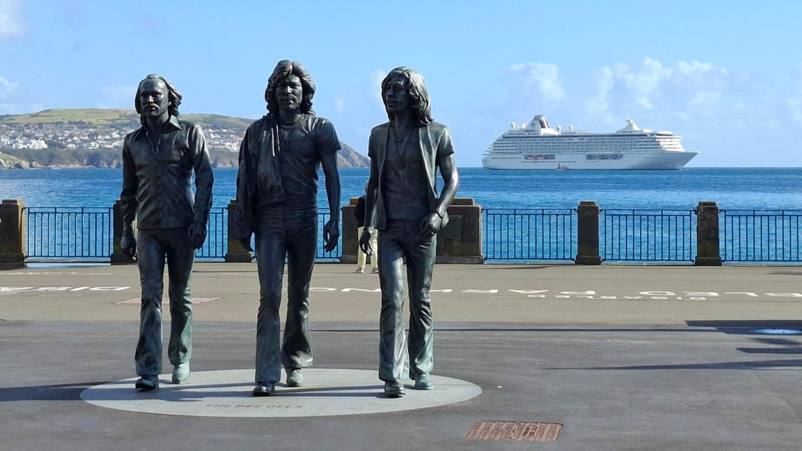 A cruise ship in Douglas bay on a sunny day can be seen in the background. A statue of the pop group The Beegees can be seen in the foreground on a concrete walkway.