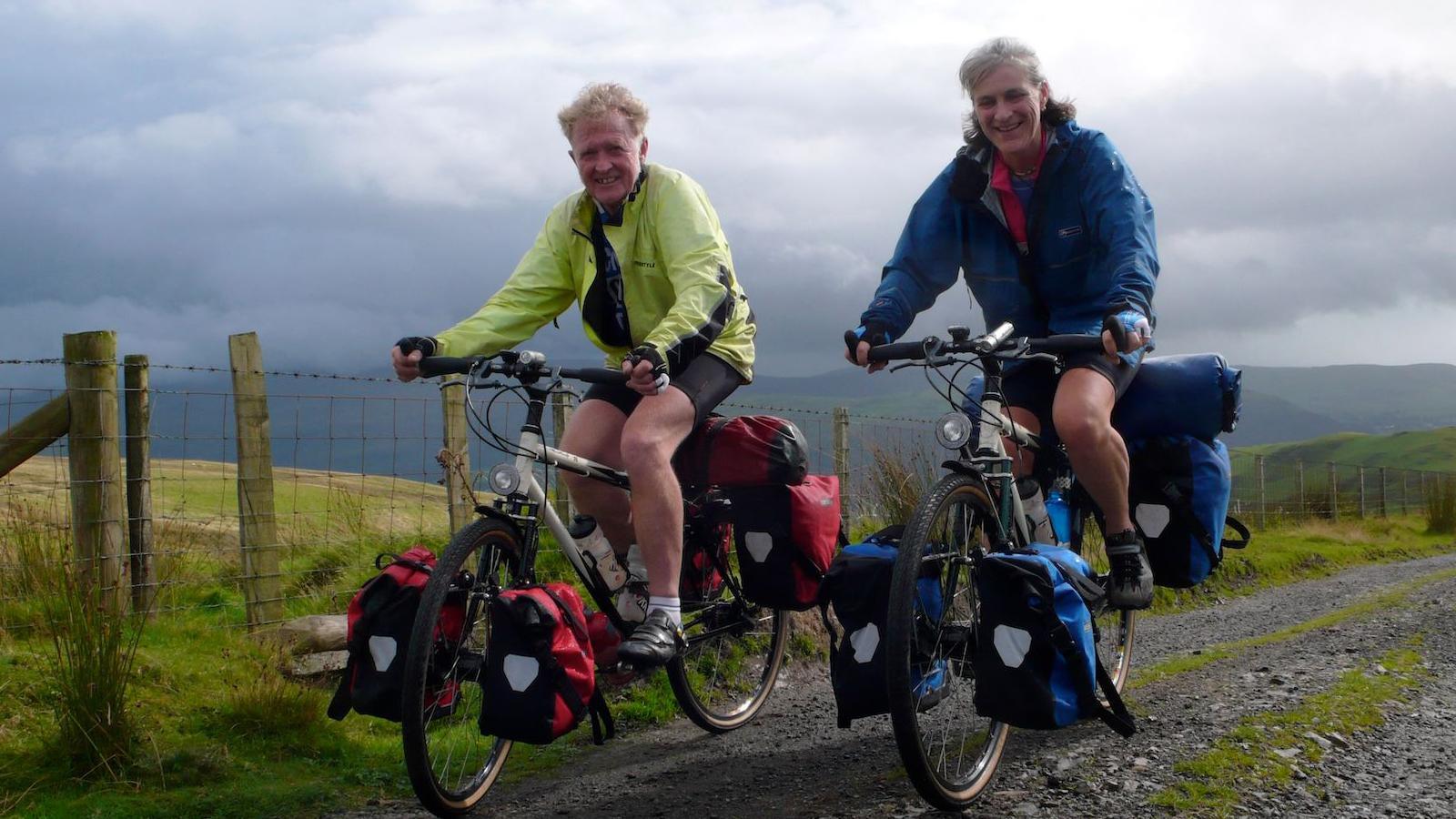 Fiona, on the right-hand side, is cycling on a bike covered in panniers next to her partner, Andy Blance, on the same set up, on a gravel path.