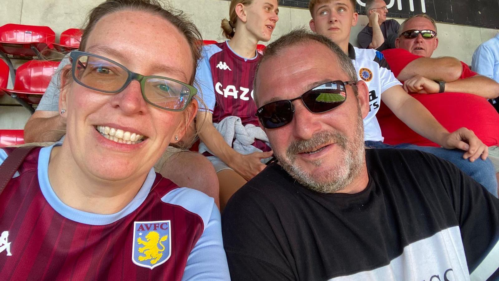 A woman in a football shirt sat a football stadium stall with red chairs next to a man in a black T-shirt. with a white stripe across the front. She has green-rimmed glasses and short dark hair, pulled back and wears a Villa shirt of claret and blue with the Villa crest on it. Behind her are other fans sat in the stadium.