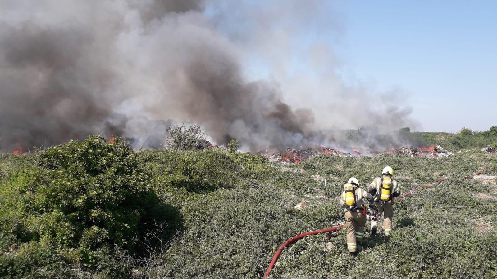 Two firefighters at Arnolds Field walking across an upward slope of green scrubland. The fire is smouldering in the background with plumes of grey and smoke is coming from the rubbish.