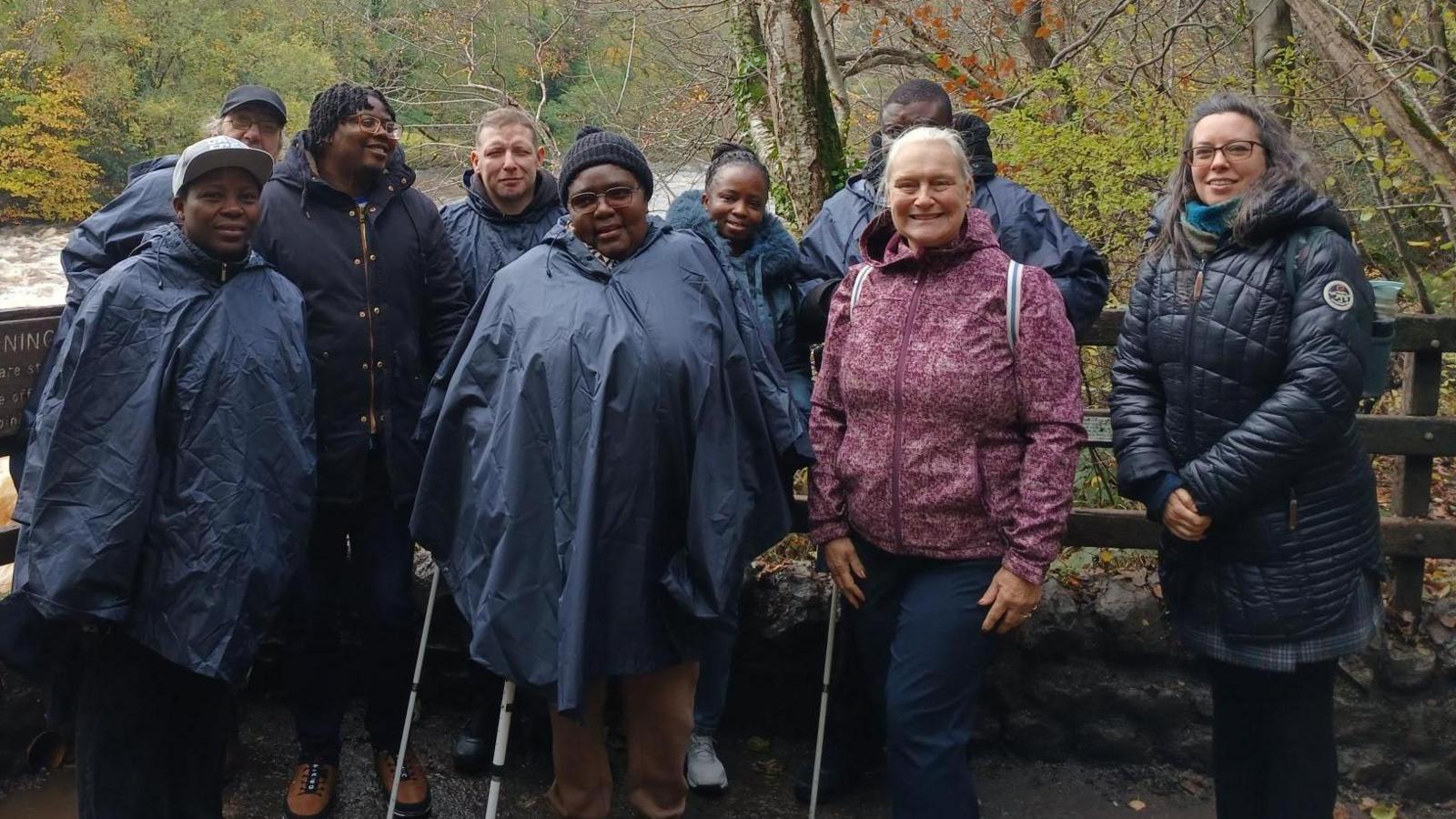 A mixed group of people stand in front of a fence by a river, posing for a picture. They all wear waterproof coats and winter clothing.
