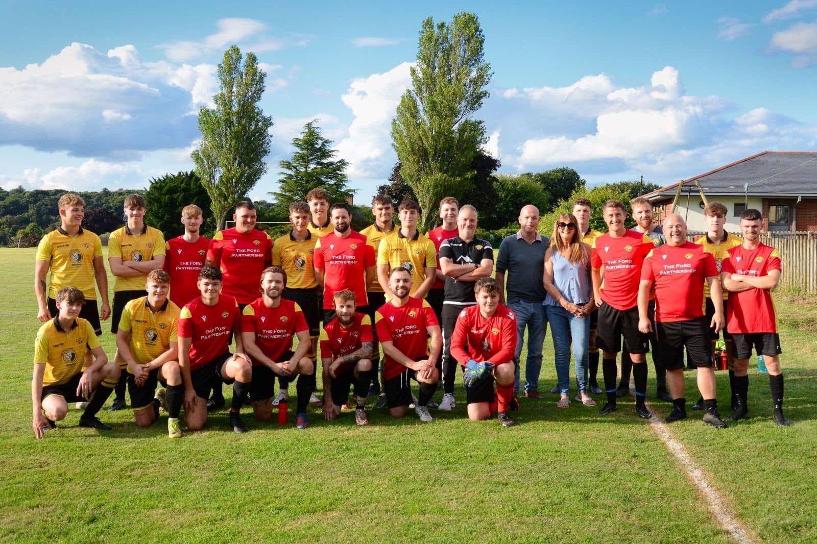 A men's football team dressed in yellow and red football kit tops and black shorts, lined up for a photo on a grass pitch, with 24 players featuring in the shot, with a referee dressed in black